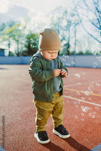 Obraz na płótnie Cheerful 2 year old boy playing outside in summer in a green jacket with soap bubbles. Toddler boy running outside and playing with bubbles. w salonie