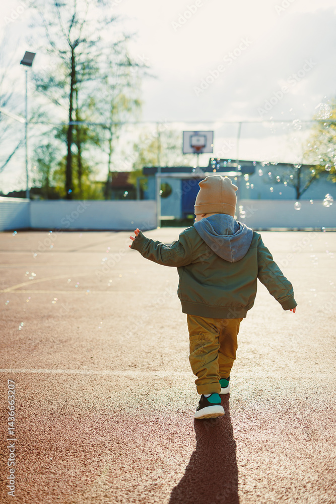Cheerful 2 year old boy playing outside in summer in a green jacket with soap bubbles. Toddler boy running outside and playing with bubbles.