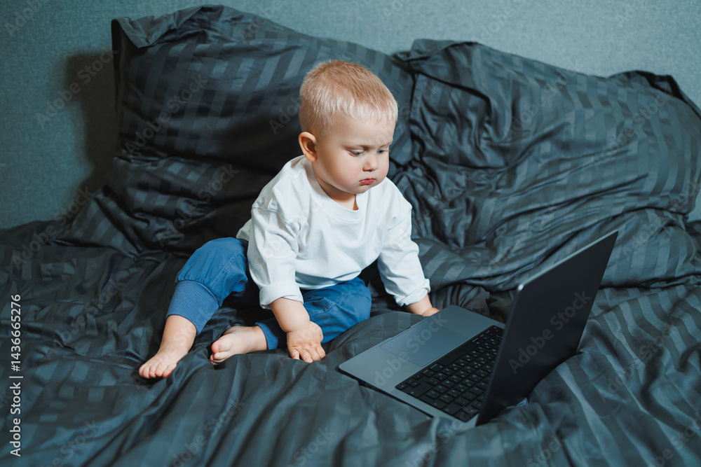 A 2-year-old boy sits in bed and presses buttons on a laptop. The child plays with a laptop.