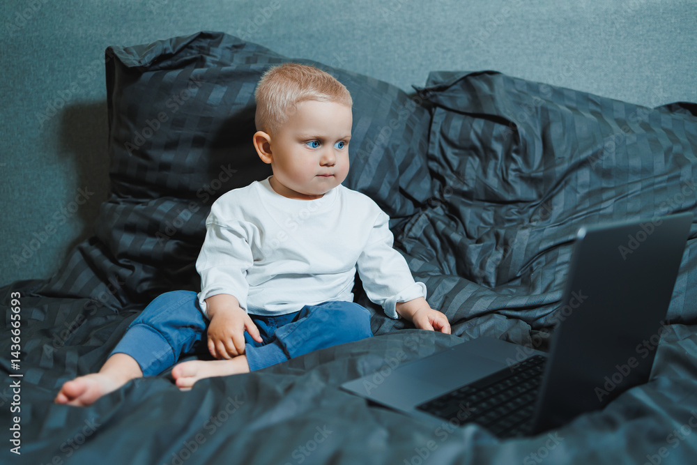 A 2-year-old boy sits in bed and presses buttons on a laptop. The child plays with a laptop.