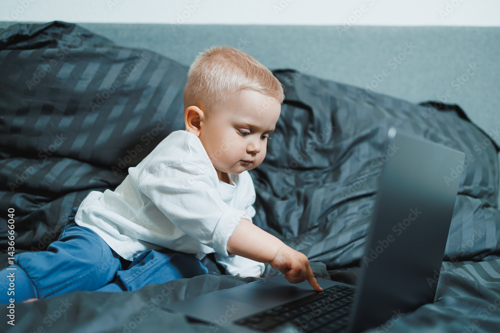 A 2-year-old boy sits in bed and presses buttons on a laptop. The child plays with a laptop.