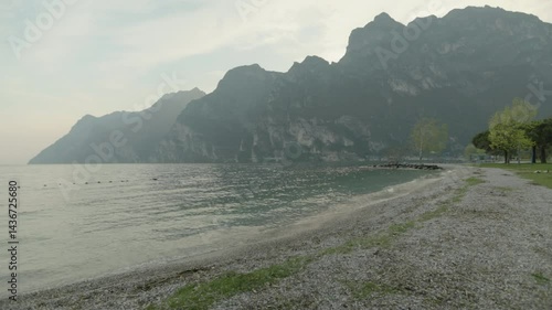 Wallpaper Mural A left to right panning shot of the bay of Riva del Garda in Italy on a slightly overcast day with water and the pebbles on the shore in the foreground and hazy majestic mountains in the background Torontodigital.ca
