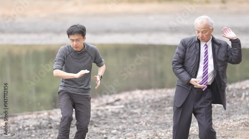 In a bamboo grove and pond park, a man in his 70s in a suit and a man in his 30s practice tai chi. Their slow movements are captured in a slow-motion video against the peaceful natural background.