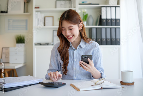 Smiling businesswoman calculating expenses and using smartphone at desk.