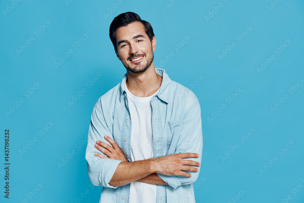 Smiling young man with crossed arms looking at the camera, wearing light blue shirt over white t-shirt, isolated on bright blue background. People lifestyle concept