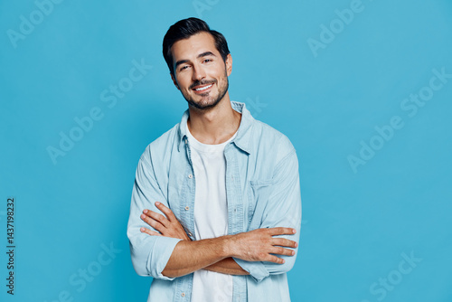 Obraz na płótnie Smiling young man with crossed arms looking at the camera, wearing light blue shirt over white t-shirt, isolated on bright blue background. People lifestyle concept w salonie