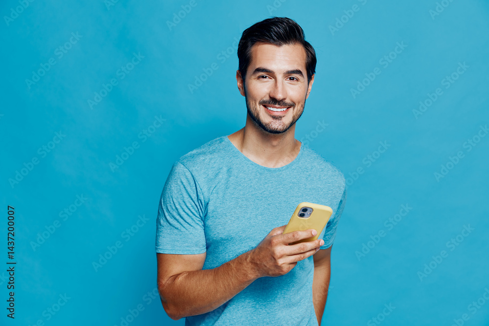 Smiling man holding a mobile phone, wearing a casual blue t-shirt, stands against a bright blue background. Cheerful mood, lifestyle theme, studio portrait.