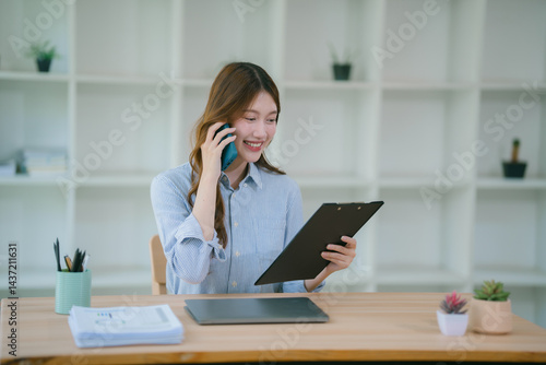Smiling asian businesswoman talking on phone and using laptop while working at office des