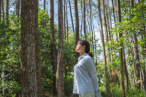 A young asian woman wearing a long-sleeve collared shirt and denim jeans pants is standing and meditating in a lush pine forest in the morning.