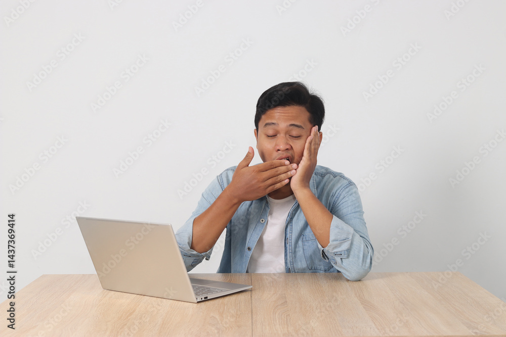 Young Asian man feeling sleepy while working on his laptop, covering his mouth as he yawns. Showing signs of fatigue and tiredness at his desk. Concept of work exhaustion and lack of energy Young Asian man feeling sleepy while working on his laptop, covering his mouth as he yawns. Showing signs of fatigue and tiredness at his desk. Concept of work exhaustion and lack of energy