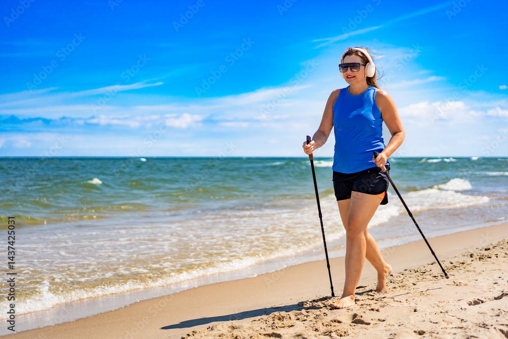Playing sports on beach in summer. Pretty mature woman practicing Nordic walking and listening to music in headphones on sandy beach on sunny day. Side view