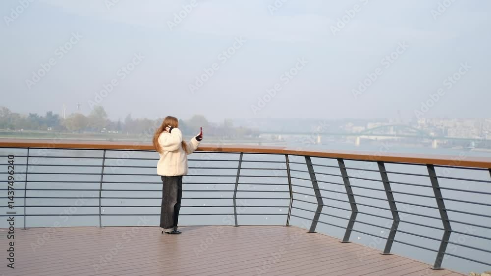 Happy girl taking a selfie outdoors on a sunny day, smiling brightly. She is wearing a cozy fleece jacket and standing by the river with city buildings in the background Happy girl taking a selfie outdoors on a sunny day, smiling brightly. She is wearing a cozy fleece jacket and standing by the river with city buildings in the background