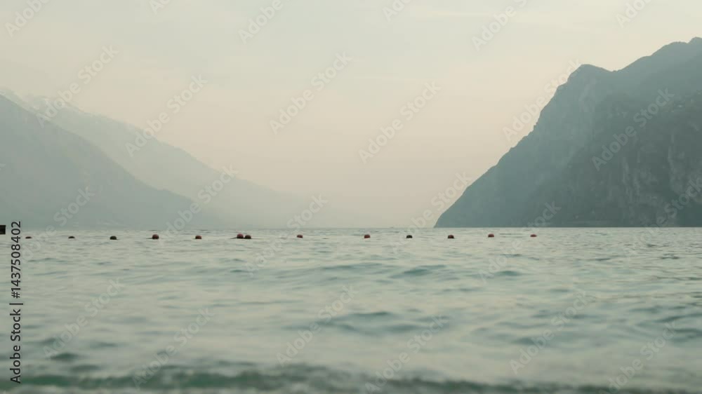 A low angle shot of Lago di Garda from the bay of Riva del Garda in Italy with hazy large mountains in the distance on an overcast day in Spring A low angle shot of Lago di Garda from the bay of Riva del Garda in Italy with hazy large mountains in the distance on an overcast day in Spring