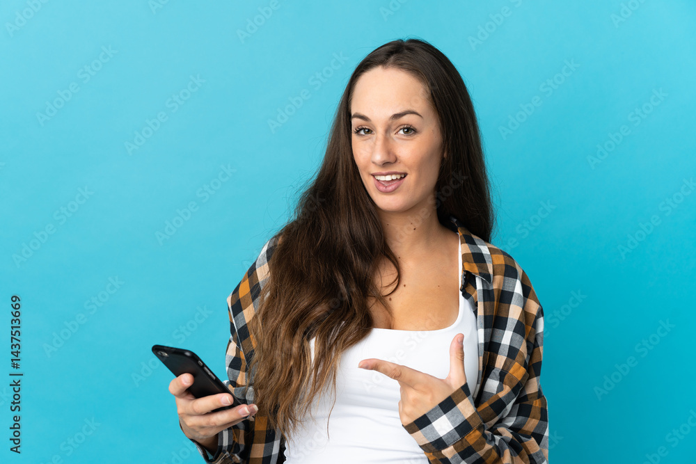 Naklejka premium Young hispanic woman over isolated blue background using mobile phone and pointing it