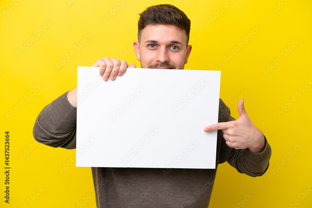 Young caucasian man isolated on yellow background holding an empty placard and hiding behind it Young caucasian man isolated on yellow background holding an empty placard and hiding behind it