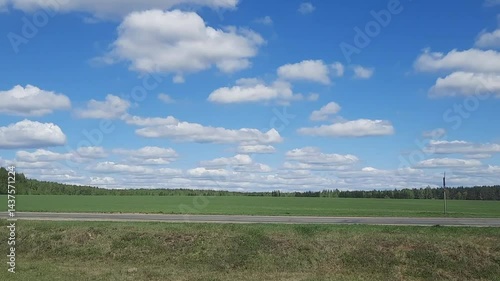 Bright blue sky with many white clouds over fields and forest. Rural landscape with green fields and forest on the horizon. Asphalt road in the foreground.