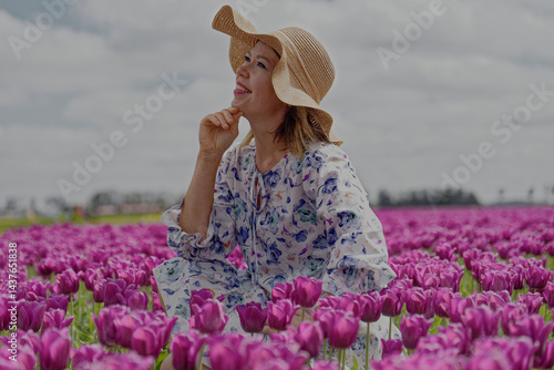 Na zdjęciu widzimy osobę w polu żółtych tulipanów, co tworzy malowniczy, wiosenny krajobraz. Ubrana w lekką, kwiecistą sukienkę, trzyma w ręku słomkowy kapelusz, dodając całości uroku i beztroski