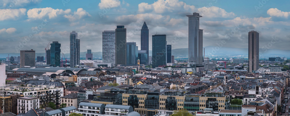 Aerial view of Frankfurt, Germany, highlighting the Messeturm, Tower 185, and Hauptbahnhof. Modern and traditional buildings with hills in the distance.
