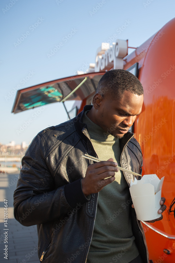 African american Man eating noodles from a takeaway box on the street. Concept of food culture, casual lifestyle and city exploration.