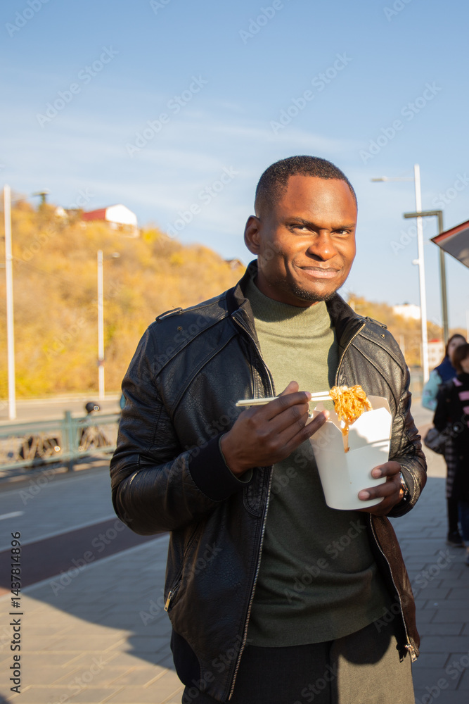 African american Man eating noodles from a takeaway box on the street. Concept of food culture, casual lifestyle and city exploration.