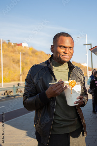 Obraz na płótnie African american Man eating noodles from a takeaway box on the street. Concept of food culture, casual lifestyle and city exploration. w salonie
