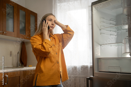 Frustrated woman in orange shirt talking on mobile phone, gesturing towards empty refrigerator in bright kitchen, reporting malfunction to customer service