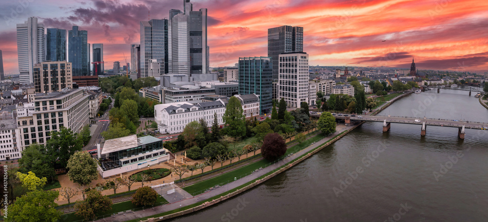 Aerial view of Frankfurt, Germany, at sunset featuring the Commerzbank Tower, Main River, a bridge, riverside greenery, and Frankfurt Cathedral spire.