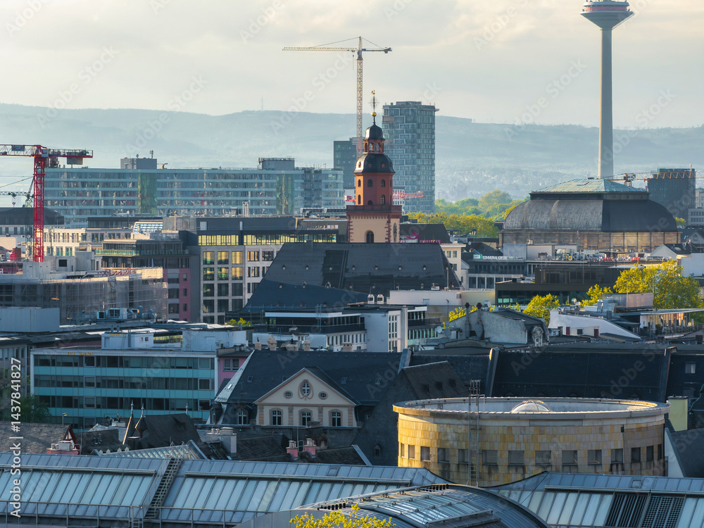 Aerial view of Frankfurt, Germany, showing Katharinenkirche, Europaturm, modern high rises, construction cranes, and green trees under soft daylight.
