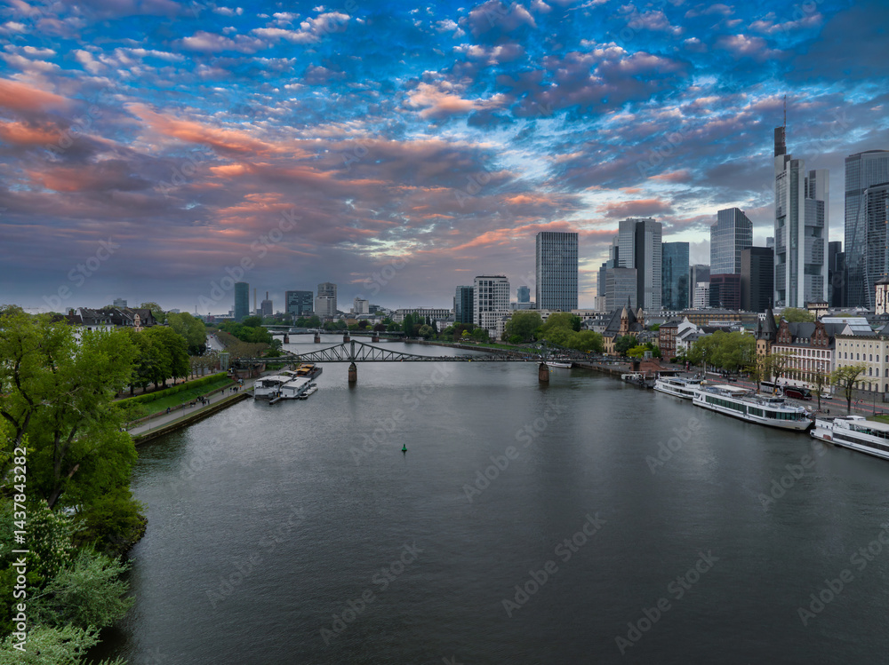 Aerial view of Frankfurt, Germany, featuring the Main River, Commerzbank Tower, Eiserner Steg bridge, and a vibrant sunset with colorful skies.