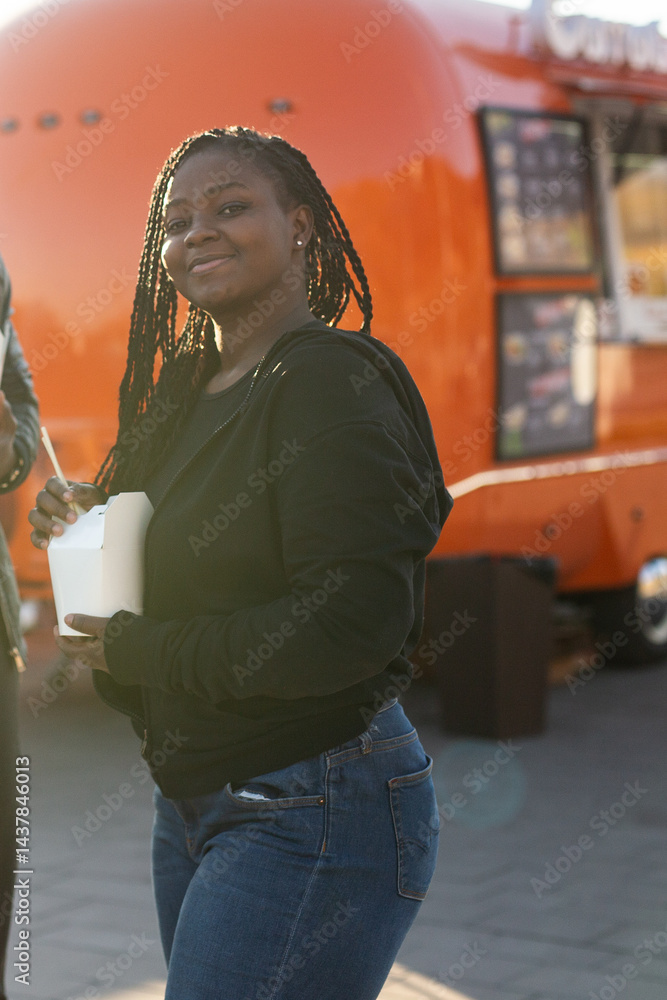 Young african american woman smiling and holding a takeaway food box near a food truck. Relaxed street food scene with warm afternoon sunlight.