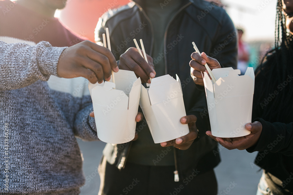 Three african american friends enjoying Asian street food outdoors on a sunny day, using chopsticks and sharing casual moments near a bright orange food truck. Warm backlight creates a cozy, friendly