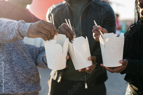 Obraz na płótnie Three african american friends enjoying Asian street food outdoors on a sunny day, using chopsticks and sharing casual moments near a bright orange food truck. Warm backlight creates a cozy, friendly w salonie