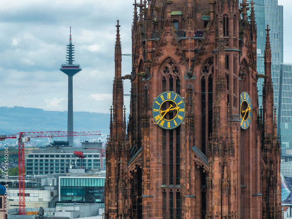 The Frankfurt Cathedral's Gothic spires and clock face stand out, with the Europaturm in the background. Cranes and modern buildings complete the urban scene.