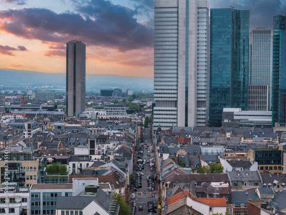 Aerial view of Frankfurt, Germany, featuring modern skyscrapers like the Deutsche Bank Twin Towers and traditional rooftops under a vibrant sunset sky.