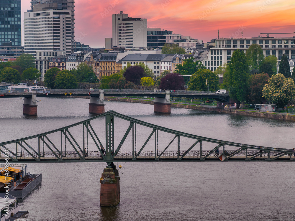 Aerial view of Frankfurt, Germany, showcasing Eiserner Steg over the Main River, modern skyscrapers, historic buildings, and a vibrant sunset sky.
