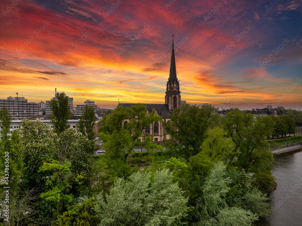 Aerial view of Frankfurt during sunset, featuring a historic church with a tall spire, lush green trees, a river, and modern city buildings in the background.