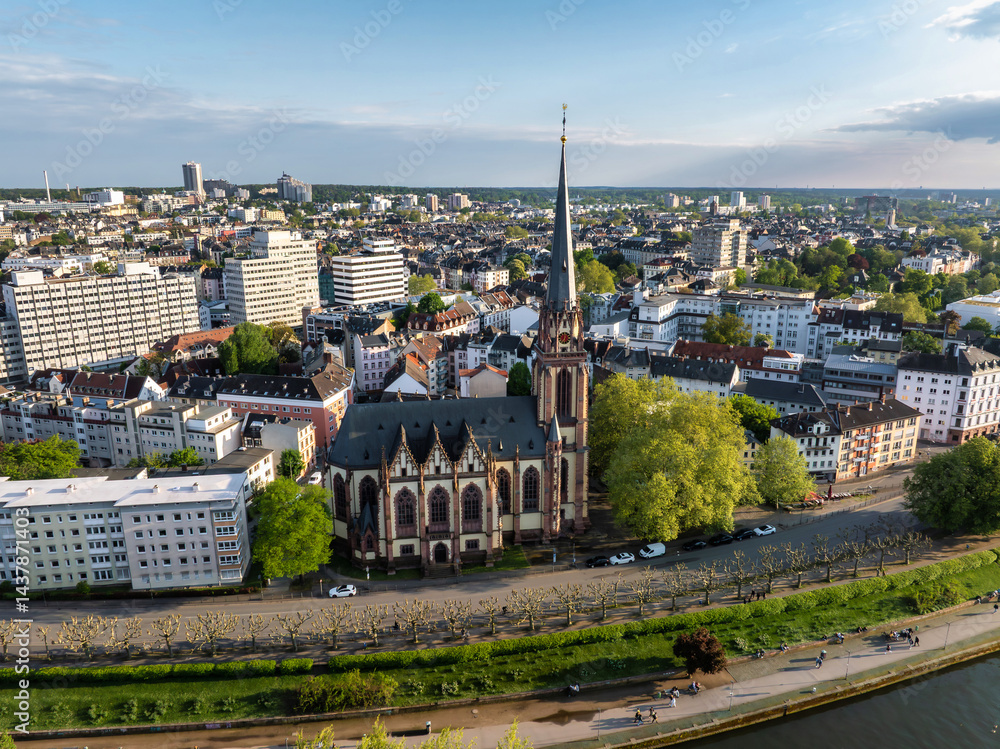 Aerial view of Frankfurt, Germany, highlighting Dreikonigskirche's Gothic spire, the Main River, tree lined pathways, and an expansive urban skyline.