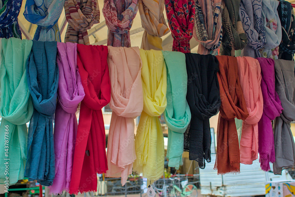 Fototapeta premium Scarves in every color are displayed neatly in a busy market. The soft fabrics hang in gentle folds, inviting touch. Bright hues create a cheerful and lively scene for shoppers.