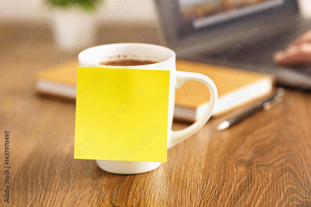 Blank yellow sticky note on coffee cup standing on wooden desk with laptop and pen in blurred background workplace organization and planning concept