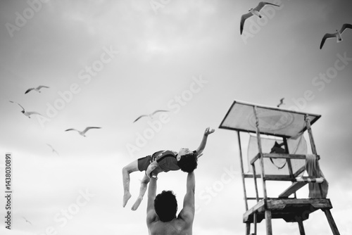 Black and white photo of father lifting child at the beach near lifeguard tower with seagulls flying overhead.