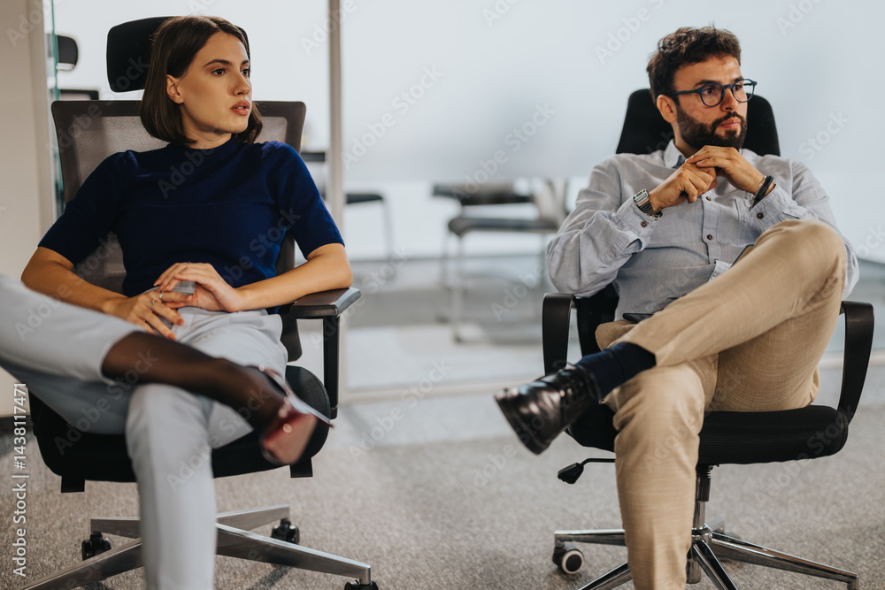 Obraz premium A man and a woman in business attire are seated in an office. They appear engaged in a serious discussion, reflecting a professional work environment. The office setting is modern and minimalistic.