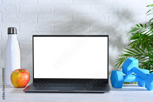 Laptop with white screen on table with dumbbells, bottle and apple