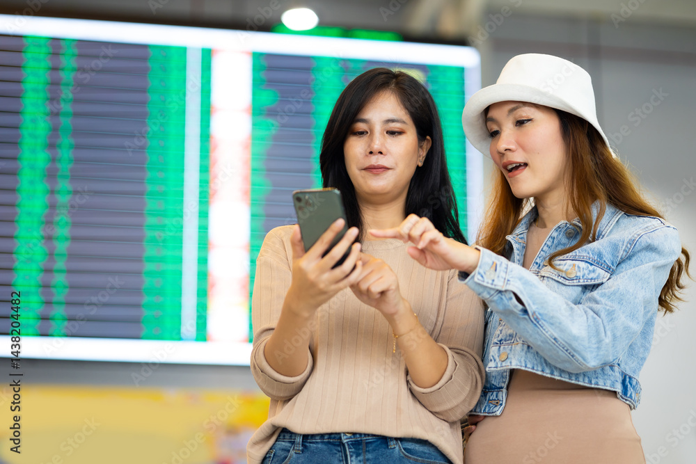 Check flight schedule on board with information display. arrival and departure board. Two Asian woman use smartphone cheking flight for travel in long weekend