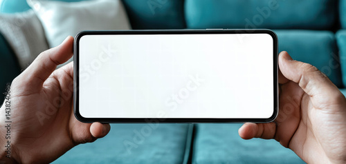 Person holding mobile device with blank screen, showcasing minimalist design. background features cozy couch, creating relaxed atmosphere