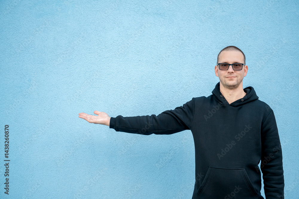 Stylish guy in glasses and black sweatshirt isolated on plain bright blue background, pointing with hand to side empty space for table or graph. Photo of model on wall background with empty space