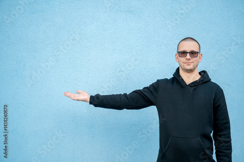 Stylish guy in glasses and black sweatshirt isolated on plain bright blue background, pointing with hand to side empty space for table or graph. Photo of model on wall background with empty space 