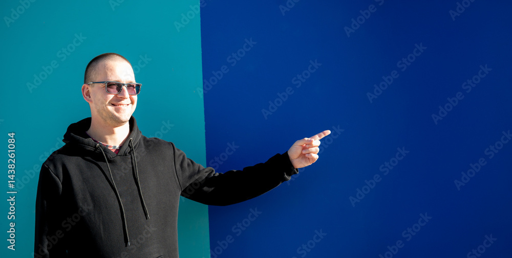 Stylish guy in glasses and black sweatshirt isolated on plain bright blue background, pointing with hand to side empty space for table or graph. Photo of model on wall background with empty space