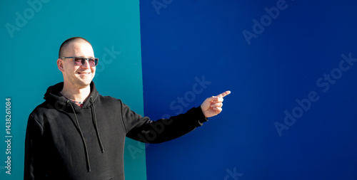 Obraz na płótnie Stylish guy in glasses and black sweatshirt isolated on plain bright blue background, pointing with hand to side empty space for table or graph. Photo of model on wall background with empty space w salonie