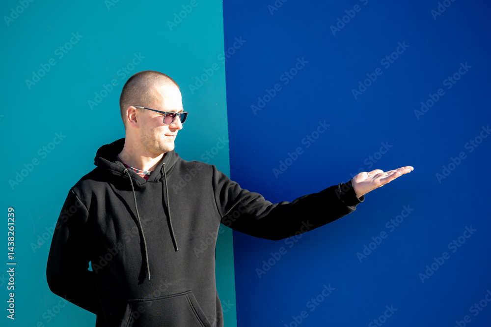 Stylish guy in glasses and black sweatshirt isolated on plain bright blue background, pointing with hand to side empty space for table or graph. Photo of model on wall background with empty space