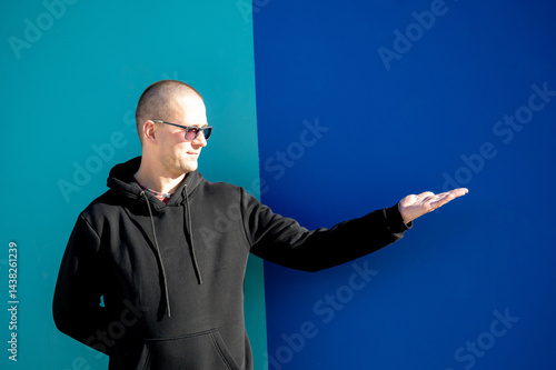 Obraz na płótnie Stylish guy in glasses and black sweatshirt isolated on plain bright blue background, pointing with hand to side empty space for table or graph. Photo of model on wall background with empty space w salonie